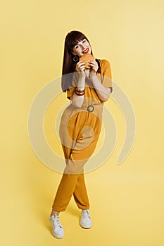 Full length portrait of happy young attractive woman in yellow overalls, standing with legs crossed, enjoying unhealthy