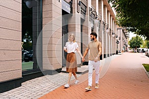 Full length portrait of happy young couple in love walking holding hands on city street. Stylish bearded man and blonde