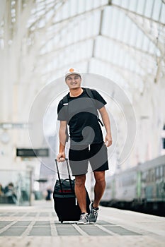 Full length portrait of a happy young man walking with suitcase at train station. Travel concept