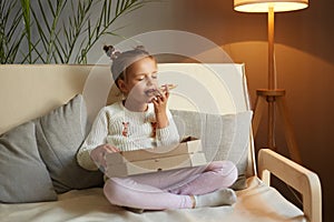 Full length portrait of hungry adorable female kid sitting on cough with crossed legs and eating fast food, holding slice of pizza