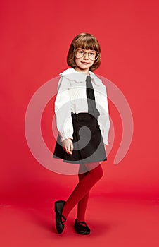 Full-length portrait of little school girl in black shirt, white blouse and glasses standing against red studio