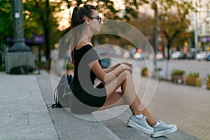 Full length portrait. Lonely girl sits on the stairs in the city, in a black dress with sunglasses, looks in profile.