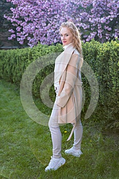 Full-length portrait of middle-aged woman in white coat with long blonde hair in spring park. Vertical frame