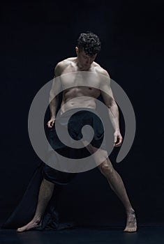 Full-length portrait of muscular, shirtless, handsome man posing in dark fabric against black studio background. Statue