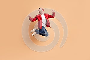Full length portrait of nice young man headphones raise fists jump wear red shirt isolated on beige color background