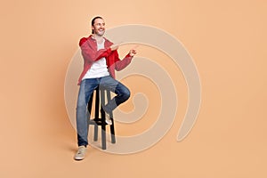 Full length portrait of nice young man sit chair direct fingers empty space wear red shirt  on beige color