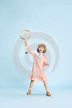 Full-length portrait of playful, happy, smiling little girl in pink dress posing with tennis racket against blue studio