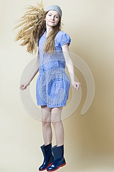 Full Length Portrait of Positive Caucasian Teenager Girl In Blue Dress and Wellington Boots Wearing Winter Hat Moving in High Jump