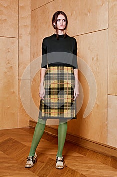 Full length portrait of a pretty brunette girl wearing a black shirt and plaid skirt and in green tights. Standing pose