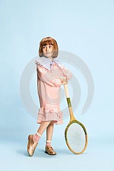 Full-length portrait of pretty little girl in pink dress posing with tennis racket against blue studio background