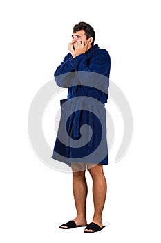 Full length portrait of scared and worried young man messy hair, wears blue bathrobe, waking up after dreamt a nightmare, bites