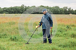 Full length portrait of senior man with metal detector and shovel searching coins or artifacts in green meadow, male enjoying