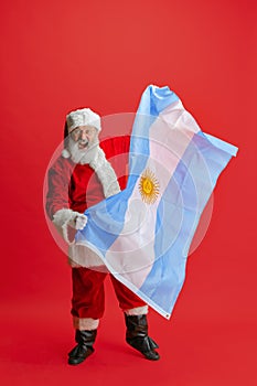 Full-length portrait of senior man wearing Santa Claus costume, holding flag of Uruguay  over red background