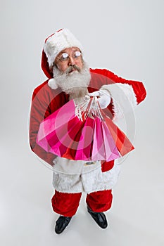 Full-length portrait of senior man wearing Santa Claus costume, holding many shopping packages isolated over white