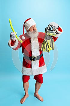 Full-length portrait of senior man wearing Santa Claus costume, holiding diving equipment mask and fins isolated over