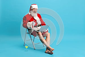 Full-length portrait of senior man wearing Santa Claus costume with laptop  over blue background. Online