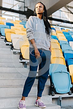Full-length portrait of the serious afro-american teenager with earphones standing on the stairs of the stadium.