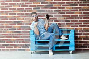 Full length portrait of shocked young bearded man in casual style and eyeglasses sitting on blue wooden pallet and pointing at