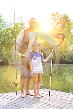 Full length portrait of smiling father and son  standing with fishing  tackles on pier against lake photo