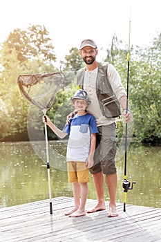 Full length portrait of smiling father and son standing with fishing tackles on pier against lake photo