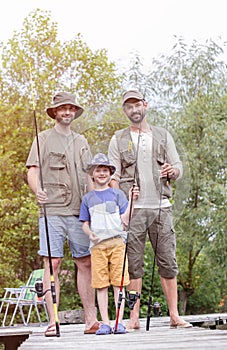 Full length portrait of smiling males standing with fishing rods on pier against trees