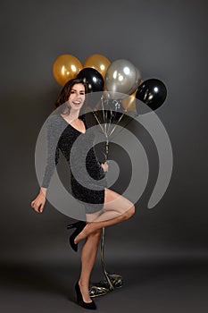 Full length portrait of a smiling stylish woman bending leg, holding balloons, celebrating new year at party, wearing sparkly