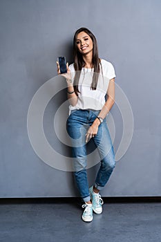 Full length portrait of a smiling woman showing blank smartphone screen isolated on a gray background.