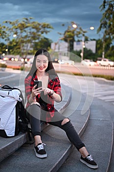 Portrait of smiling young woman sitting on stairs in the city and using smart phone.