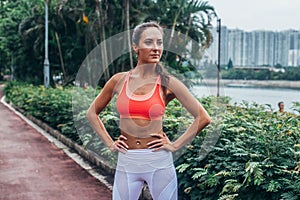 Full-length portrait of sporty fit model standing in park posing in pink sports bra and white leggings with hands on