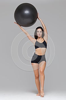 Full length portrait of sporty woman in black sportswear holding an exercise ball above her head isolated on grey