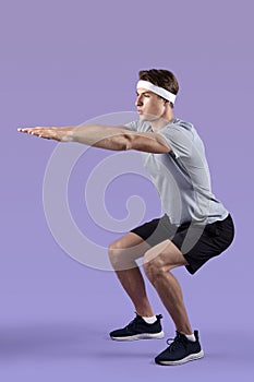 Full length portrait of sporty young guy doing squats on violet studio background