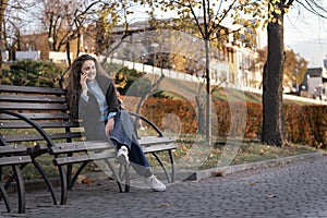 Full-length portrait of student girl sitting on bench in park talking on phone and laughing. Young woman uses smartphone