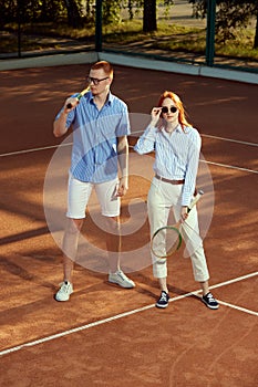 Full-length portrait of stylish, beautiful young man and woman in casual clothes posing on opendoor tennis court on warm