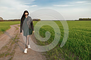 Full length portrait of a stylish girl walking along a green field. A young smiling woman is walking in nature. Green spring