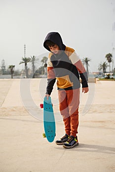 Full length portrait of stylish schoolboy in sportswear, holding skateboard, standing on an outdoor skatepark playground