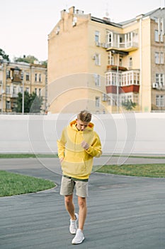 Full-length portrait of stylish young man in yellow hoodie posing at camera on background of velodrome and old buildings. Vertical