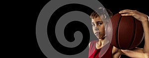 Full-length portrait of teen boy, basketball player in red uniform with ball posing isolated over black background