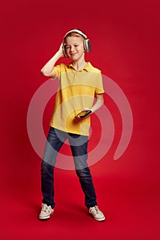 Full-length portrait of teenager, dancing while listening music in headphones against red studio background. Little DJ.