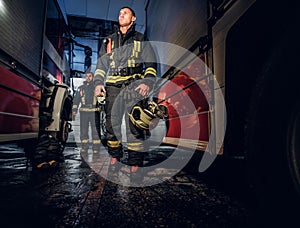 Full-length portrait of two brave firemen in protective uniform walking between two fire engines in the garage of the