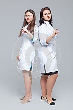 Full-length portrait of two female doctors standing back-to-back hold ready-to-inject syringes