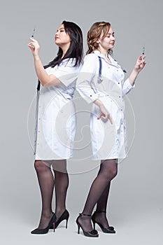 Full-length portrait of two female doctors standing back-to-back hold ready-to-inject syringes