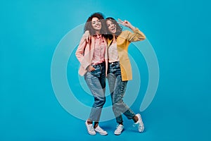 Full length portrait of two stylish girls. Indoor photo of young women with brunette hair isolated on blue background