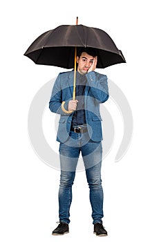 Full length portrait of upset and thoughtful businessman standing under open umbrella keeps palm to face looking down confused