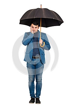 Full length portrait of upset and thoughtful businessman standing under open umbrella keeps palm to face looking down confused