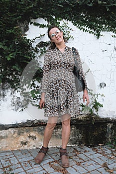 Full-length portrait of woman with funny glasses and floral print dress standing near the white wall with plants.
