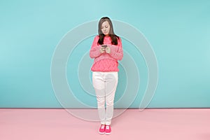 Full length portrait of woman in knitted rose sweater, white pants texting sms on cellphone isolated on bright pink blue