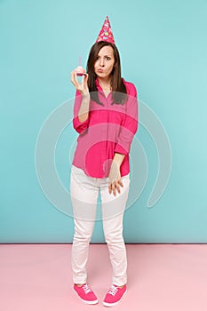 Full length portrait woman in rose shirt blouse, white pants, birthday hat with cupcake posing isolated on bright pink