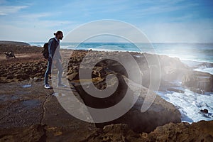 Full length portrait of a young adult man standing on the cliff by Atlantic ocean while waves breaking on the rock