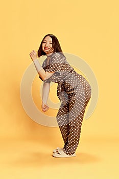Full-length portrait of young, beautiful, asian girl in silk brown homewear, pajamas and slippers dancing against yellow