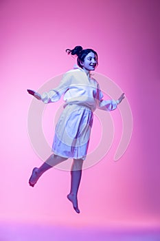 Full-length portrait of young beautiful girl in white silk outfit dancing, flying isolated on pink background in neon